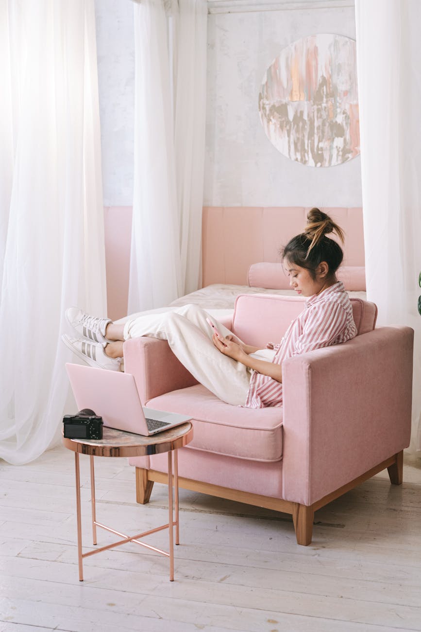 Young Asian woman using cellphone and laptop while sitting on a pink armchair indoors.
