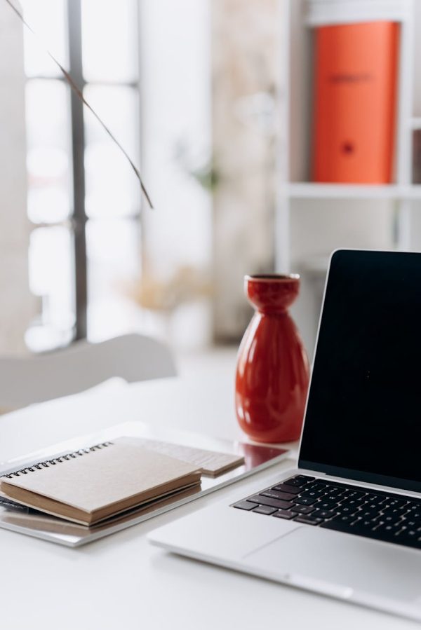 A minimalist home office setup featuring a laptop, notebook, and a red vase on a white desk.