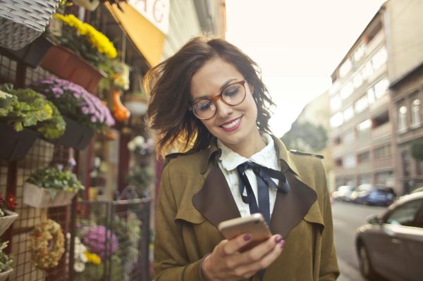 A smiling woman uses her phone outside a colorful urban flower shop on a sunny day.