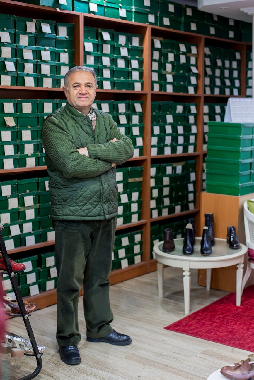Full body of calm ethnic male customer in uniform standing with crossed arms in shoe store against shelves with boxes and looking at camera