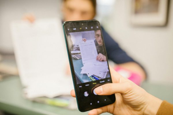 Close-up of student using smartphone to photograph examination papers in a classroom setting.
