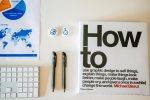 Flat lay of a creative workspace with 'How To' book, chart, pens, and keyboard in minimalist style.