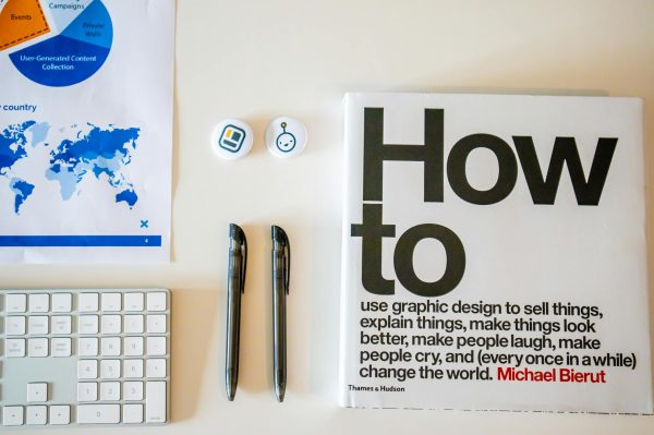 Flat lay of a creative workspace with 'How To' book, chart, pens, and keyboard in minimalist style.