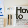 Flat lay of a creative workspace with 'How To' book, chart, pens, and keyboard in minimalist style.