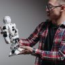 An adult man examines a humanoid robot against a gray background, exploring modern technology.